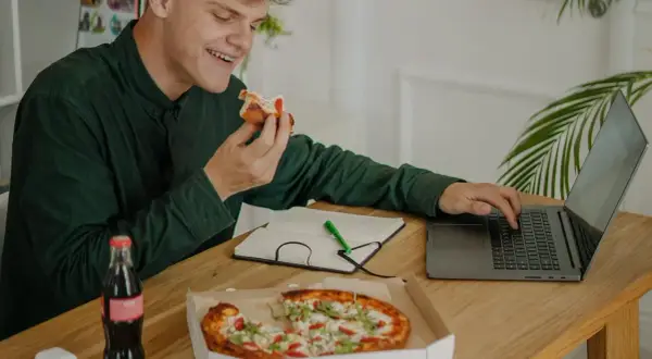 Młody mężczyzna je pizzę, pracując przy laptopie. Obok butelka napoju i notatnik. Dba o dietę, pamiętając o insulinie aspart.