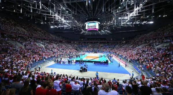 Trybuna kibiców w biało-czerwonych barwach dopingująca zawodników podczas meczu siatkówki. W tle boisko i zawodnicy.