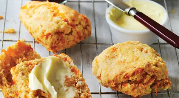 Trois scones américains dorés, dont un coupé en deux et garni de beurre, sur une grille. Un pot de confiture et du beurre à tartiner sont à proximité.