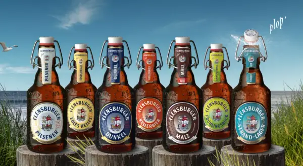 Flensburger Pils Bier Marken am Strand. Eine Möwe fliegt über das Meer, während die Flaschen mit Bügelverschluss im Gras stehen.