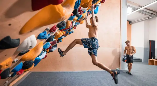 Ein Mann klettert an einer Boulderwand, während ein anderer Mann daneben steht. Der Unterschied zwischen Klettern und Bouldern wird hier deutlich: keine Seile, nur Griffe und eine Matte.