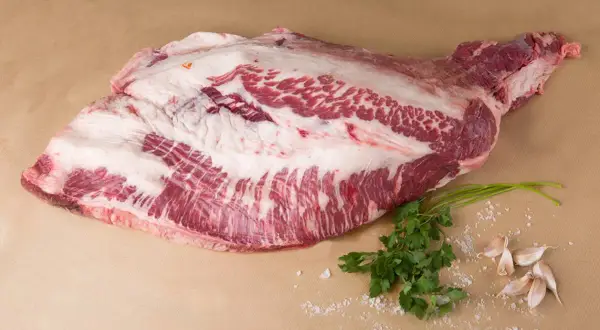 Un corte de carne cruda, con grasa marmoleada, listo para convertirse en un delicioso pastrami que es. Acompañado de perejil y ajos.