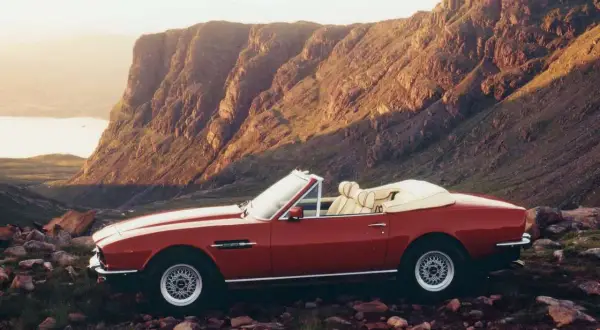 Röd Aston Martin V8 Vantage cabriolet parkerad i en stenig, bergig terräng med en sjö i bakgrunden.