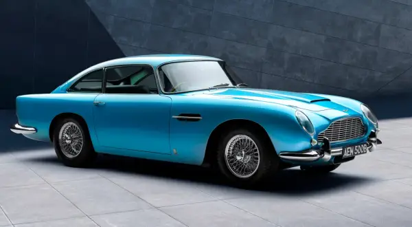 Błękitny Aston Martin DB5 z charakterystycznymi szprychowymi felgami, lśniącym chromem i elegancką linią nadwozia, zaparkowany przed nowoczesną, betonową ścianą.