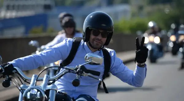 Mężczyzna w kasku i niebieskiej koszuli jedzie na motocyklu i pokazuje znak pokoju.