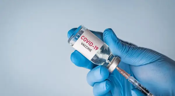 Ręka w niebieskiej rękawiczce trzyma fiolkę z napisem "COVID-19 VACCINE" i strzykawkę, gotowa do szczepienia.