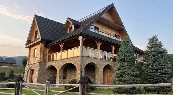 Drewniana willa z kamienną podstawą i tarasem, otoczona drzewami i ogrodzeniem.