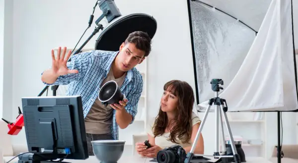 Mężczyzna, technik fotografii i multimediów, pokazuje kobiecie lampę. W tle sprzęt studyjny.