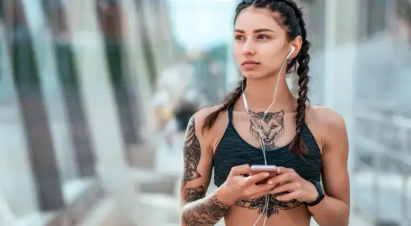Kobieta z warkoczami i słuchawkami na uszach, trzymająca telefon. Widoczny delikatny tatuaż między piersiami w kształcie kota.
