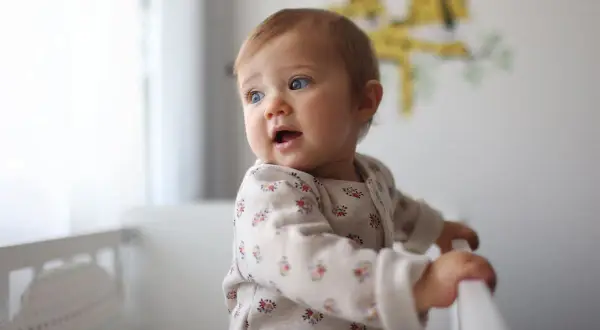 Un bébé aux yeux bleus, prénom fille, se tient dans son lit, curieux du monde.