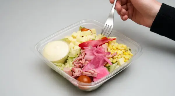 En hand tar en gaffel ur en plastlåda med pastasallad med skinka och fetaost, majs, tomat och dressing.