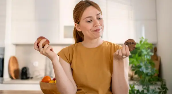 Vrouw twijfelt tussen een appel en een chocoladekoekje. De strijd tegen het verslaafd zijn aan eten is zichtbaar in haar blik.