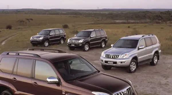 Cztery samochody Toyota Land Cruiser 120, w tym jeden brązowy, dwa czarne i jeden srebrny, stoją na polnej drodze wśród traw.