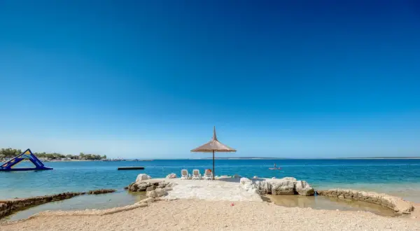 Słoneczna plaża z parasolem i leżakami w camping village Simuni. W tle wodny plac zabaw i spokojne morze.