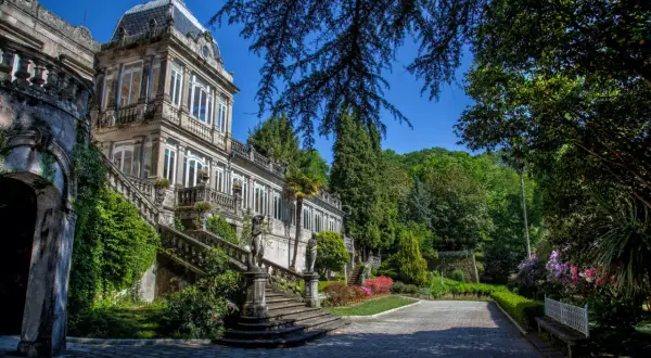El Pazo de Lourizán, una mansión señorial con escalinatas cubiertas de hiedra y un jardín exuberante, se alza majestuoso bajo un cielo azul.