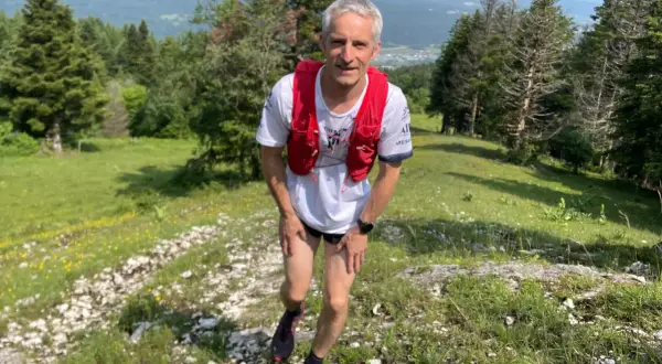 Un coureur en tenue de trail, sac à dos rouge, sur un sentier escarpé près d'Oyonnax, profitant d'un paysage verdoyant et vallonné.