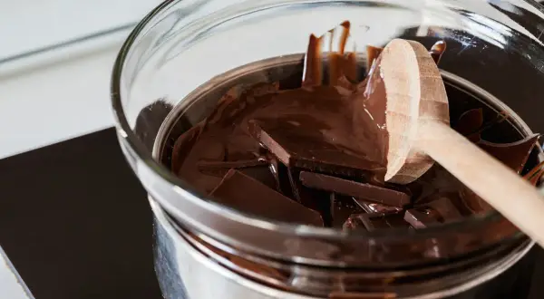 Trozo de chocolate oscuro derritiéndose en baño maría, con una cuchara de madera. Así es como derretir chocolate para postres.