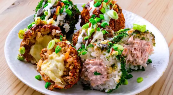 Sechs bunte Onigiri mit verschiedenen Füllungen und Toppings, umgeben von Frühlingszwiebeln und Saucen.