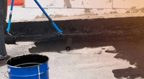 Osoba wykonuje hydroizolację piwnicy od wewnątrz, malując ścianę czarną masą bitumiczną za pomocą wałka.