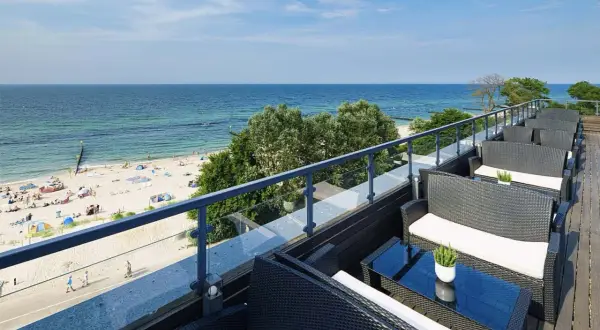 Wygodne fotele i stoliki na tarasie z widokiem na piaszczystą plażę i błękitne morze. Idealne miejsce na relaks, gdy dostępne są wolne pokoje z widokiem na morze.