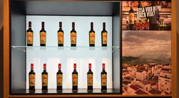 Butelki likieru Amaro Lucano ustawione na półkach, obok opisu produktu i włoskiego krajobrazu.