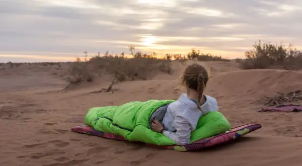 Fin du **trek dans le désert**. Une jeune femme en sac de couchage vert admire le coucher de soleil sur les dunes.