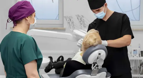 Dentysta w masce i czarnym uniformie wykonuje zabieg. Obok stoi asystentka w zielonym fartuchu. To zdjęcie szwów po zabiegu.