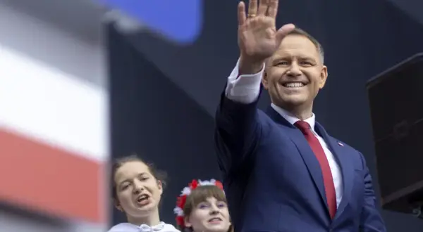Uśmiechnięty polityk w garniturze pozdrawia tłum. W tle dwie młode kobiety, jedna z wiankiem na głowie. To może być część kampanii wyborczej, pozew w trybie wyborczym.