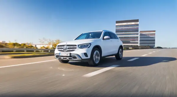Biały Mercedes GLC 200 jedzie po pustej drodze, w tle nowoczesne budynki biurowe.