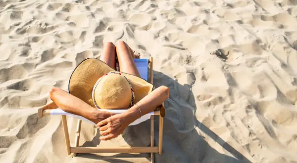 Relaks na plaży, idealne miejsce, gdzie na wakacje w lipcu. Kobieta w kapeluszu leży na leżaku wśród piaszczystych wydm.