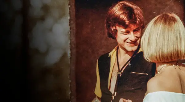 Uśmiechnięty mężczyzna w stylu lat 70. rozmawia z kobietą. Scena jak z filmu "Tulipan".
