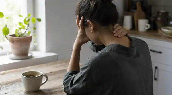Donna con mal di testa, mani sulla testa e collo, accanto a una tazza di caffè. Forse la caffeina fa male.