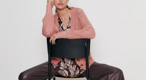 Elegancka kobieta w różowym swetrze Gerry Weber i brązowych spodniach z ekoskóry siedzi na czarnym krześle.