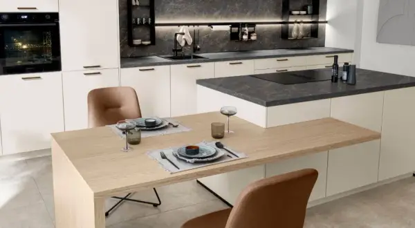 Cocina moderna con encimeras de piedra oscura y gabinetes blancos. La mesa de madera clara y las sillas de cuero marrón complementan los materiales para cocinas modernas.