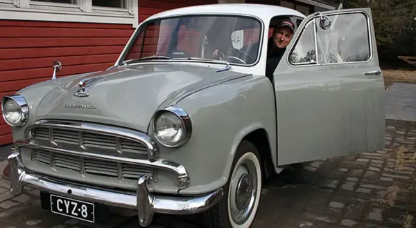 Man ler vid ratten i en grå Morris Oxford med öppen dörr.