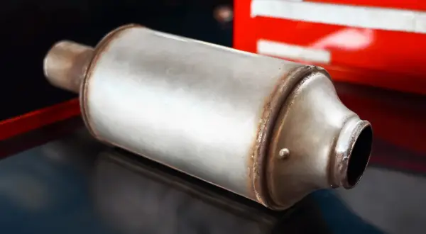 Metalowy tłumik, element układu wydechowego, prawdopodobnie z samochodu z filtrem GPF, leży na ciemnym tle.