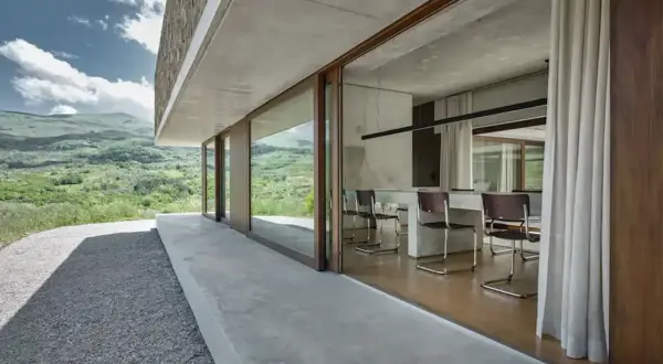 Interno moderno con tavolo e sedie, vista su paesaggio verde. Architettura in stile brutalista con cemento a vista.