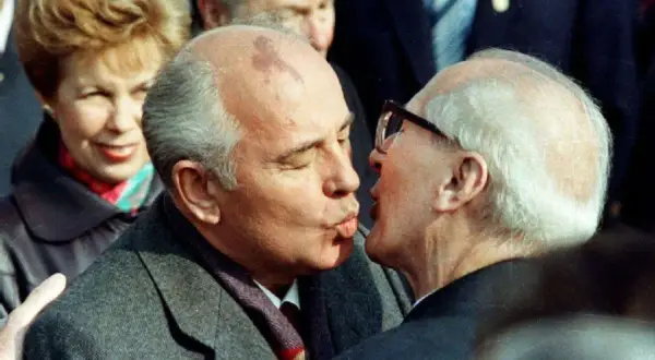 Gorbatschow und Honecker im Bruderkuss. Eine Frau im Hintergrund lächelt.