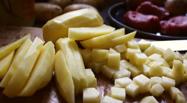 Skalade och skurna potatisar i olika former, redo för att bli härligt råstekt potatis. I bakgrunden syns hela potatisar och kött.