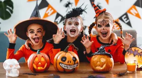 Tre barn med läskig ansiktsmålning och halloween-kostymer njuter av en halloween fest med pumpor och dekorationer.