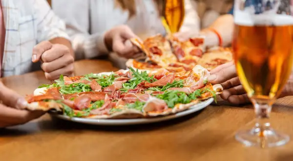 Un gruppo di amici condivide una deliziosa pizza con prosciutto e rucola, accompagnata da una fresca birra.