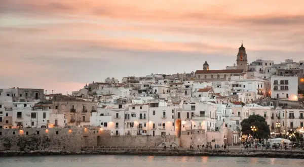Vieste lungomare al tramonto, con case bianche aggrappate alla collina e il mare calmo.