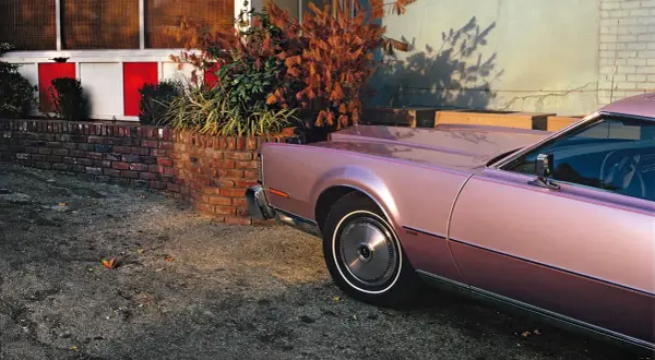 Un'iconica william eggleston foto: una Lincoln rosa metallizzato parcheggiata accanto a un muro di mattoni e un cespuglio autunnale.