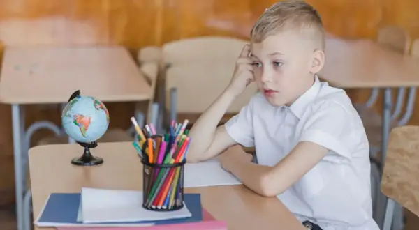 Chłopiec w białej koszuli siedzi przy biurku z globusem i kredkami. Zamyślony, może czeka na odroczenie obowiązku szkolnego.