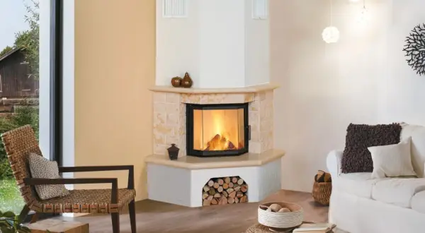 Gemütliches Wohnzimmer mit einem Eck-Kachelofen, der einladend brennt. Ein Rattansessel und ein weißes Sofa laden zum Entspannen ein.