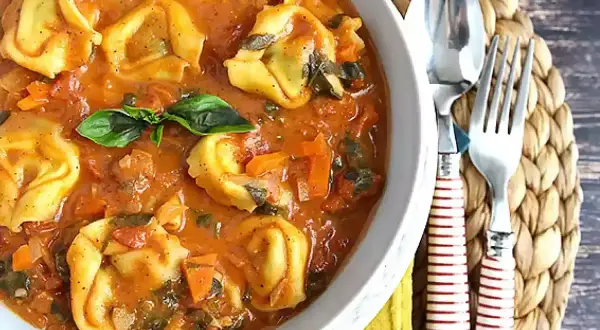 Pyszne tortellini z mięsem w kremowym sosie pomidorowym z warzywami, udekorowane listkiem bazylii. Obok widelec i łyżka.