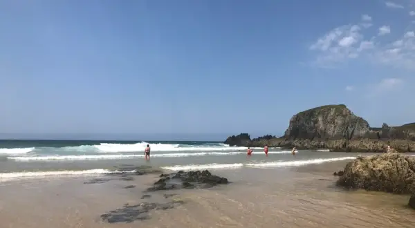 Gente disfrutando del mar en la playa de Frouxeira, con olas rompiendo y una gran roca verde al fondo bajo un cielo azul.