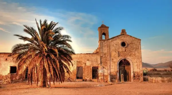 El cortijo del fraile, una edificación antigua con una palmera imponente, bajo un cielo azul y nubes al atardecer.