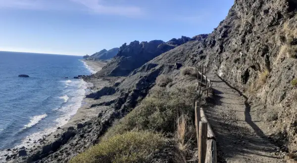 Sendero costero con vistas al mar Mediterráneo y montañas. Un camino de tierra con barandilla de madera serpentea por la ladera, ideal para un paseo marítimo en Mojácar Playa.