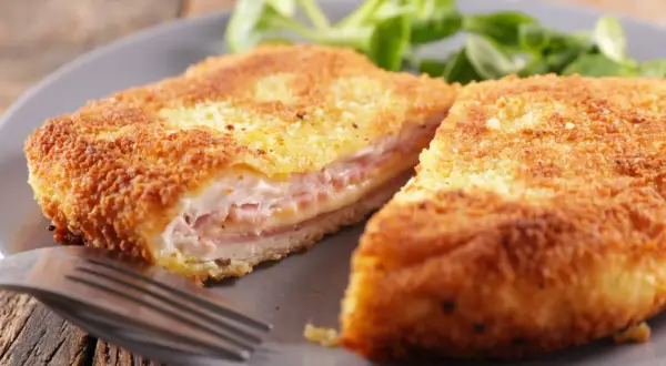 Złocisty kotlet po szwajcarsku, przekrojony na pół, odsłaniający soczyste mięso, szynkę i ser. Podany z zieloną sałatką.